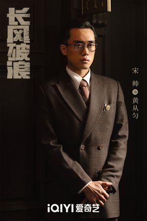 a portrait photograph of Huang Congyun, a young Chinese man with neat slicked back hair and wire rimmed glasses, wearing a brown, formal, double breasted suit with a white shirt and mid brown tie. His jacket pocket bears a small badge which marks him as an employee of the Central Bank, and there is a metallic pen visible in the pocket. His hands are clasped together in front of him and he stands straight-backed with a neutral facial expression.